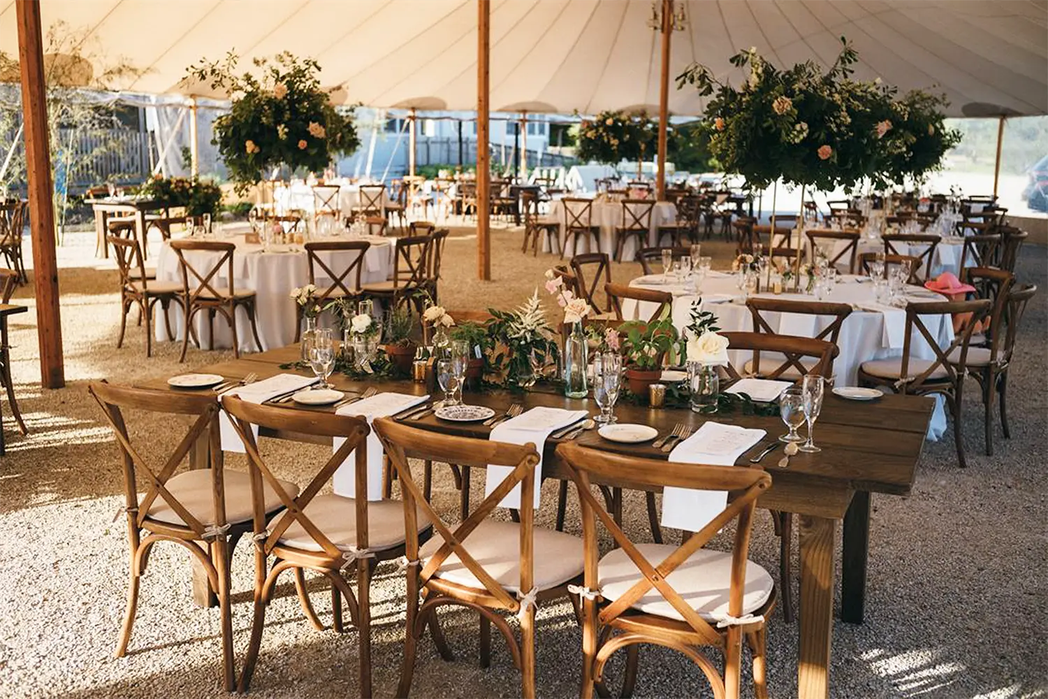 Elegant outdoor wedding reception under a white tent with wooden tables, cross-back chairs, and lush floral centerpieces.
