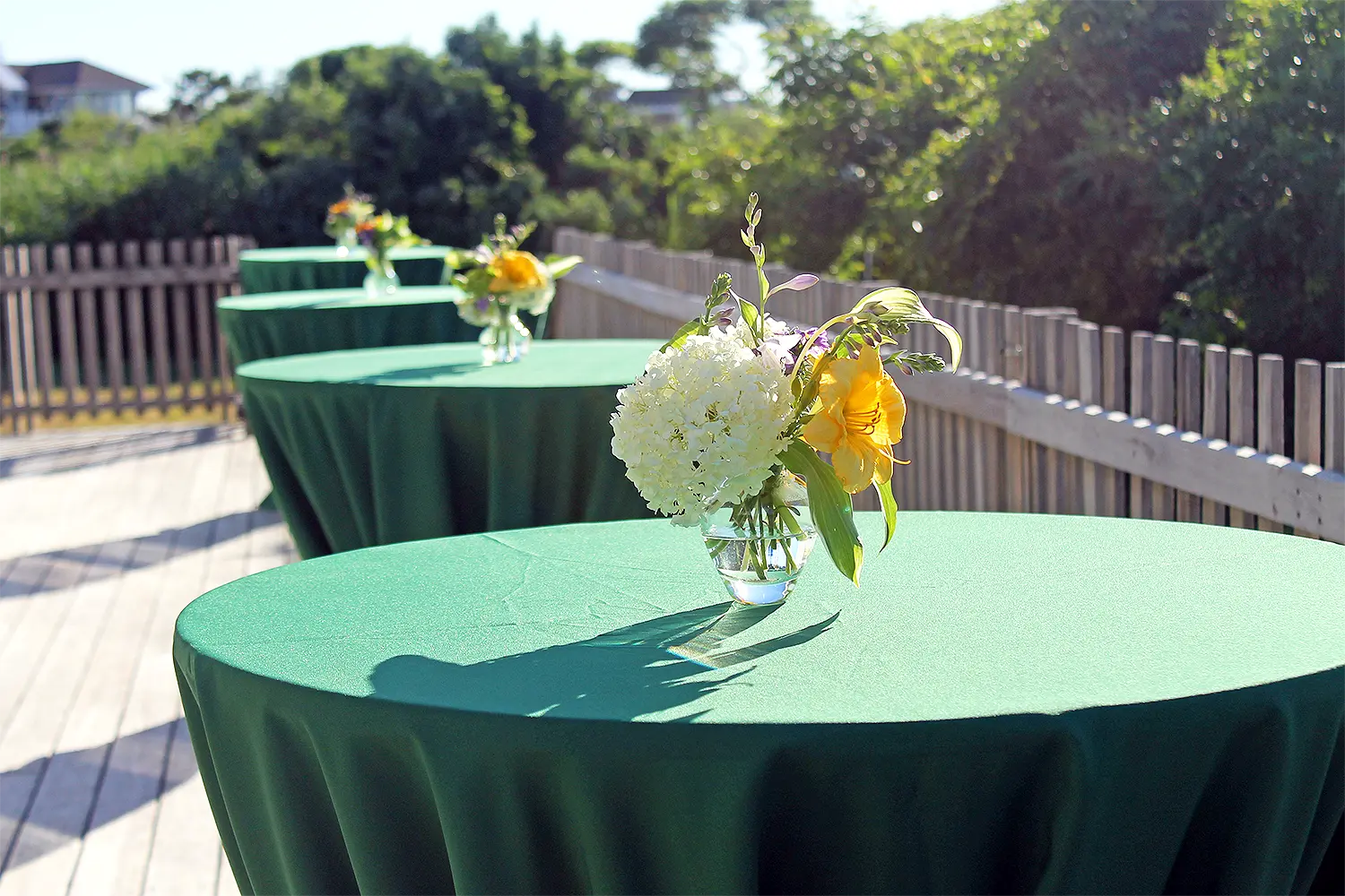 Outdoor cocktail tables with deep green linens and small floral arrangements set along a sunlit wooden deck.