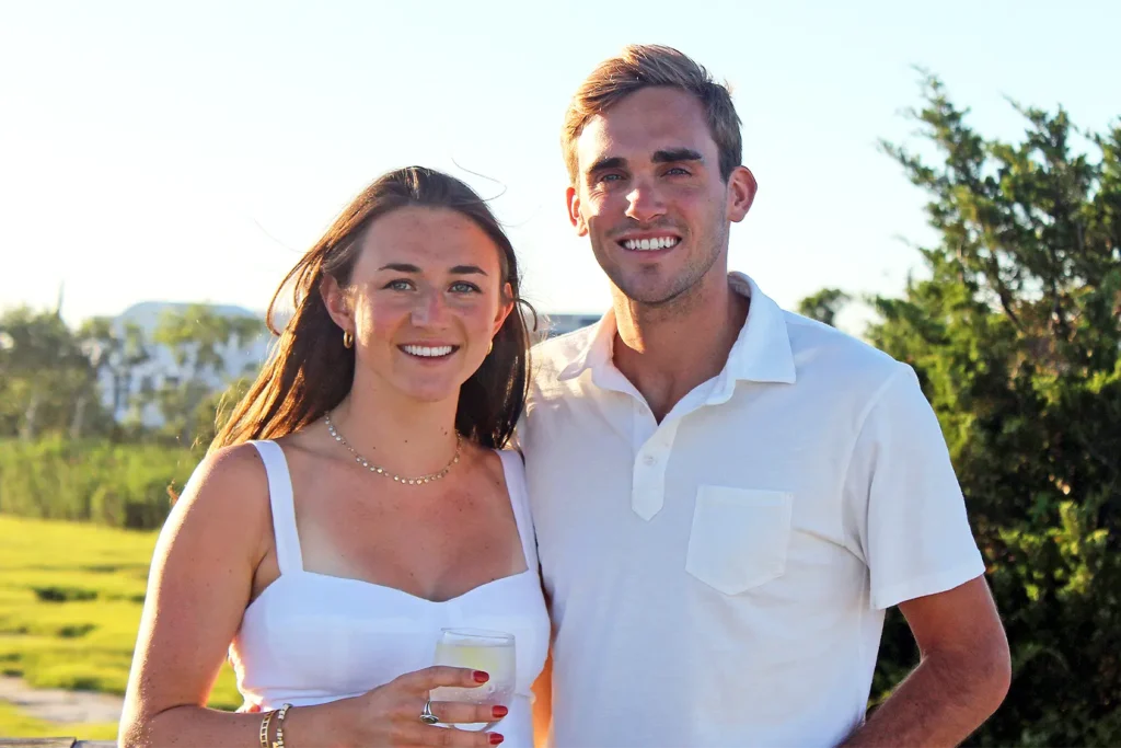 A smiling young couple stands outdoors holding drinks in a sunlit setting.