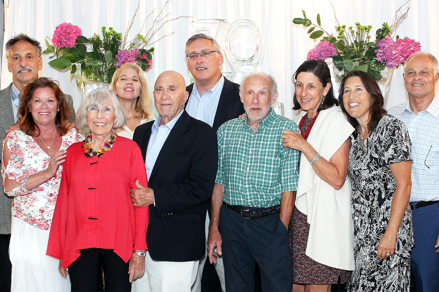 A group of older adults pose together smiling at a formal indoor celebration with floral arrangements behind them.