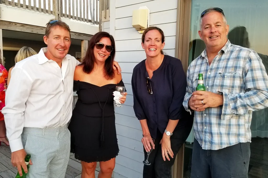 Four adults smile and pose together outdoors on a deck during a private social gathering, holding drinks.