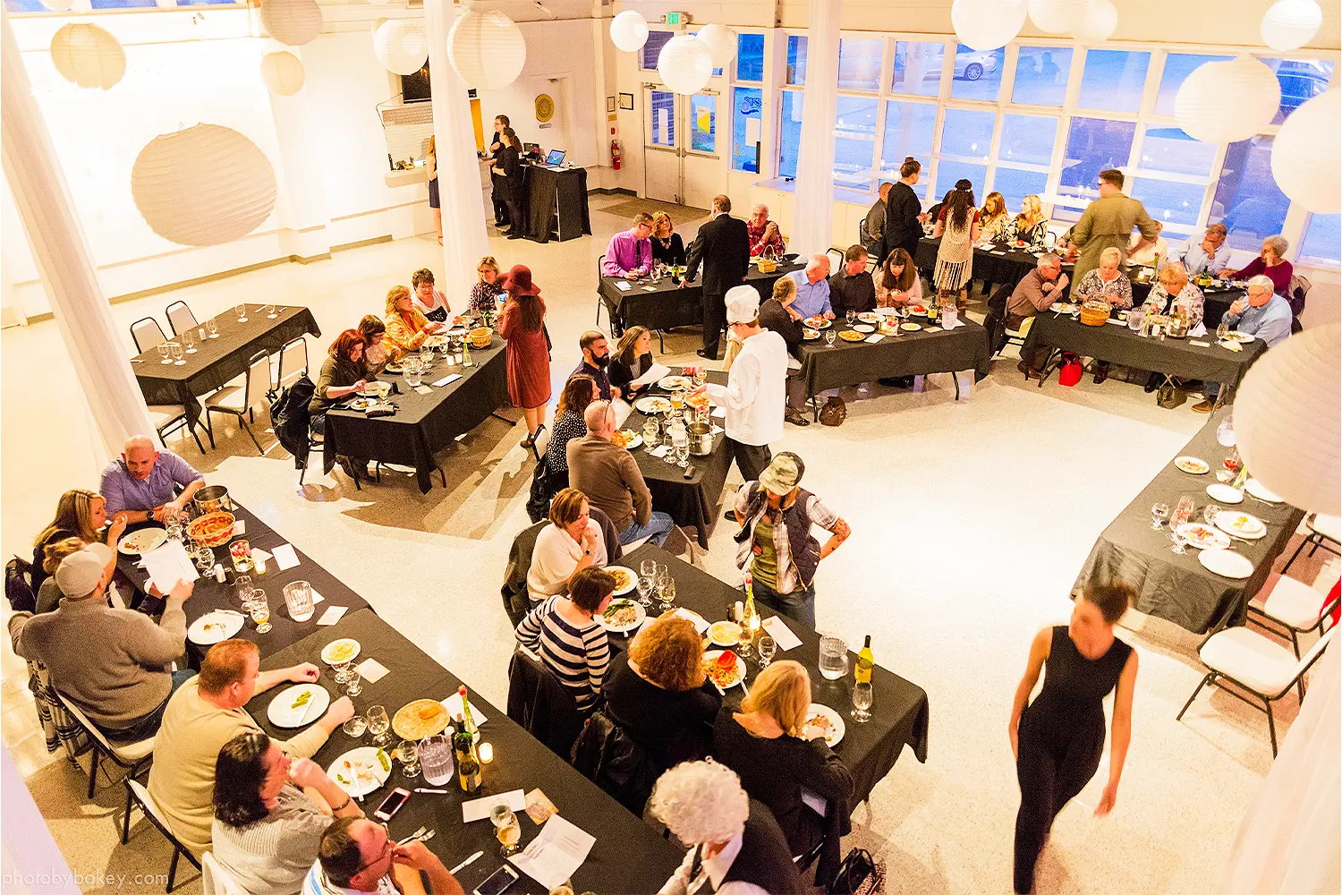 Overhead view of guests seated at long tables inside a bright event space, dining and socializing during a private indoor gathering.