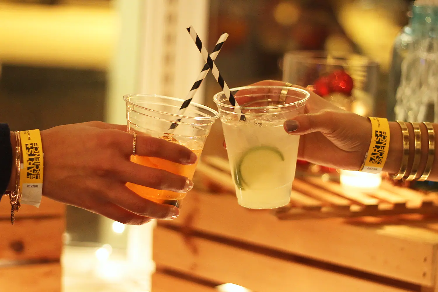 Two people clink clear plastic cups filled with cocktails, wearing yellow LBIF Arts + Science wristbands during an evening event.