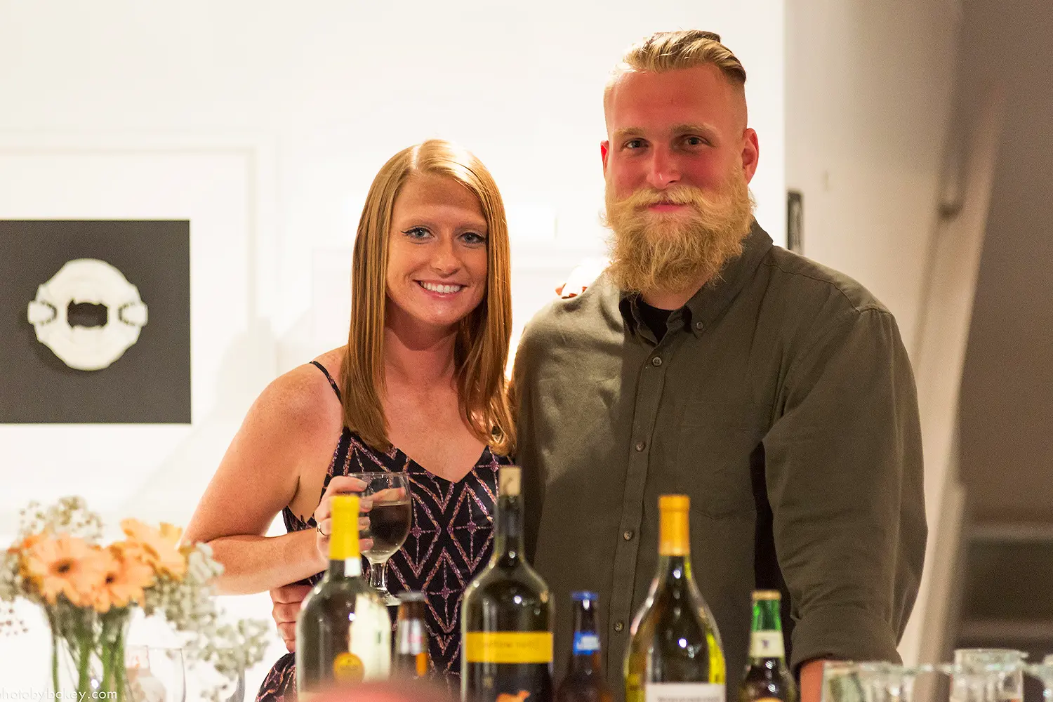 Two guests smile toward the camera while standing behind a bar with wine bottles and drinks at an indoor LBIF event.