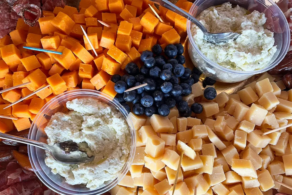 Close-up of a cheese and charcuterie spread with cubed cheddar, aged cheese, blueberries, cured meats, and small cups of creamy spread.