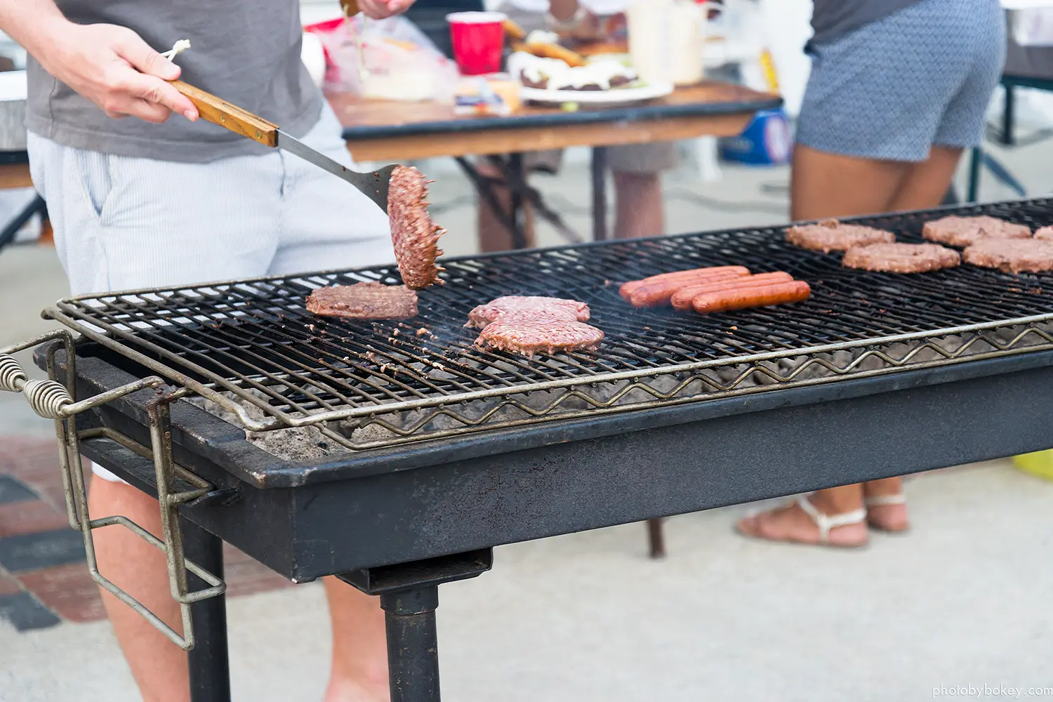Burgers and hot dogs cooking on an outdoor grill as a person flips a burger patty with a spatula.