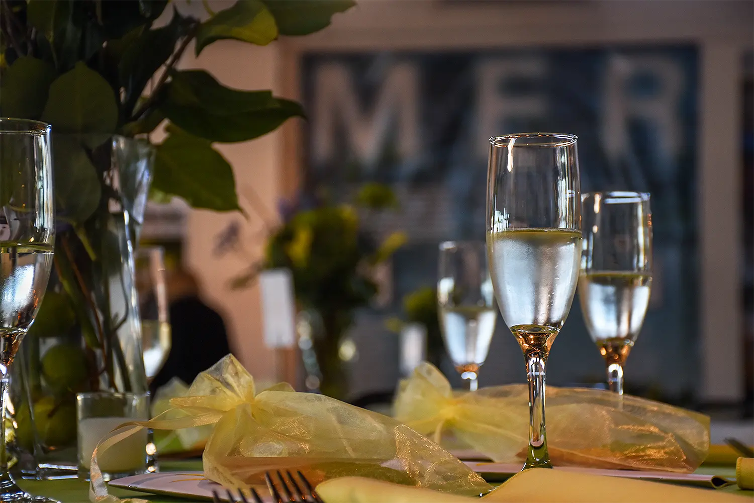 Champagne flutes filled with sparkling wine sit on an elegantly set table with soft yellow napkins and floral arrangements.
