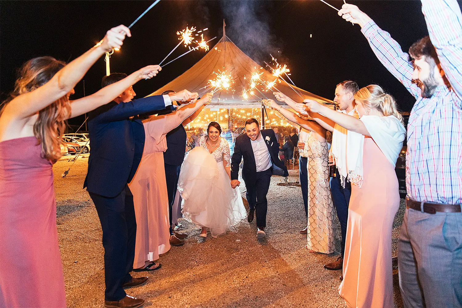 Newlyweds run hand in hand through a sparkler send-off at night as wedding guests cheer on both sides.