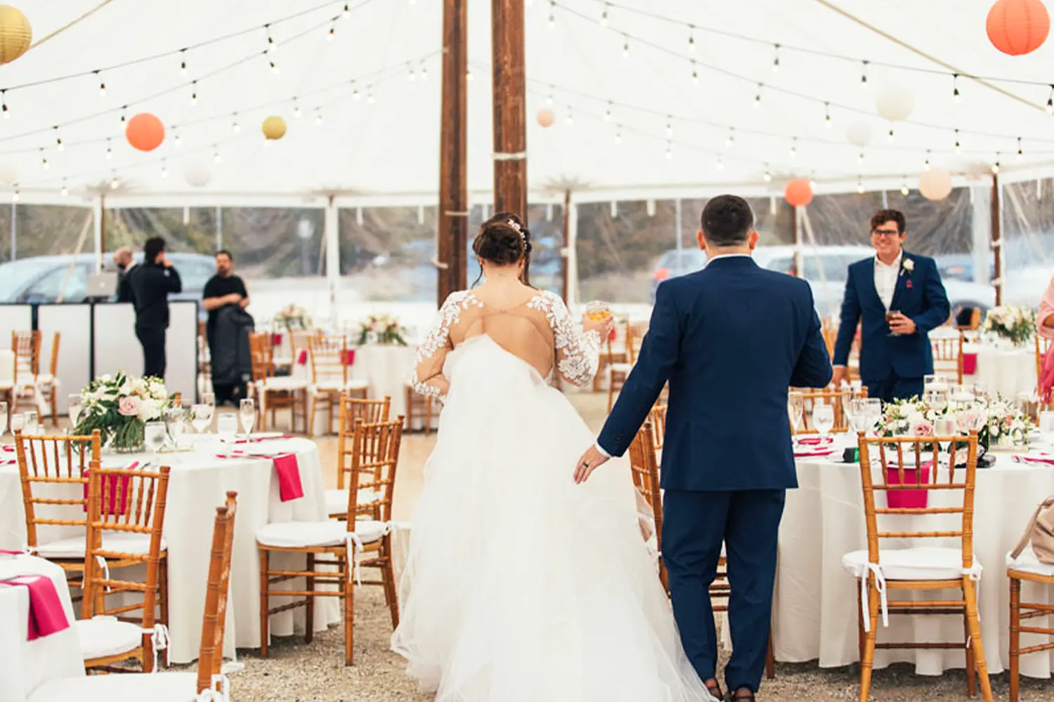 Bride and groom walk hand in hand into a wedding reception tent decorated with string lights, round tables, and wooden chairs.