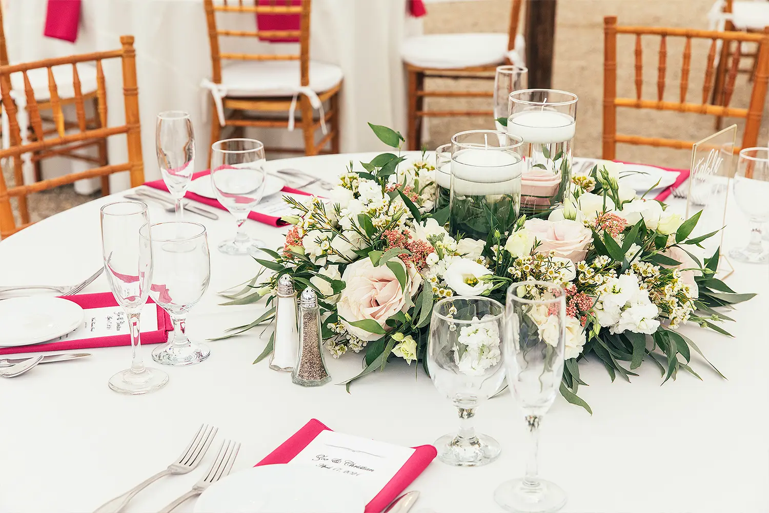 Elegant wedding reception table set with white linens, glassware, pink napkins, and a floral centerpiece with candles.
