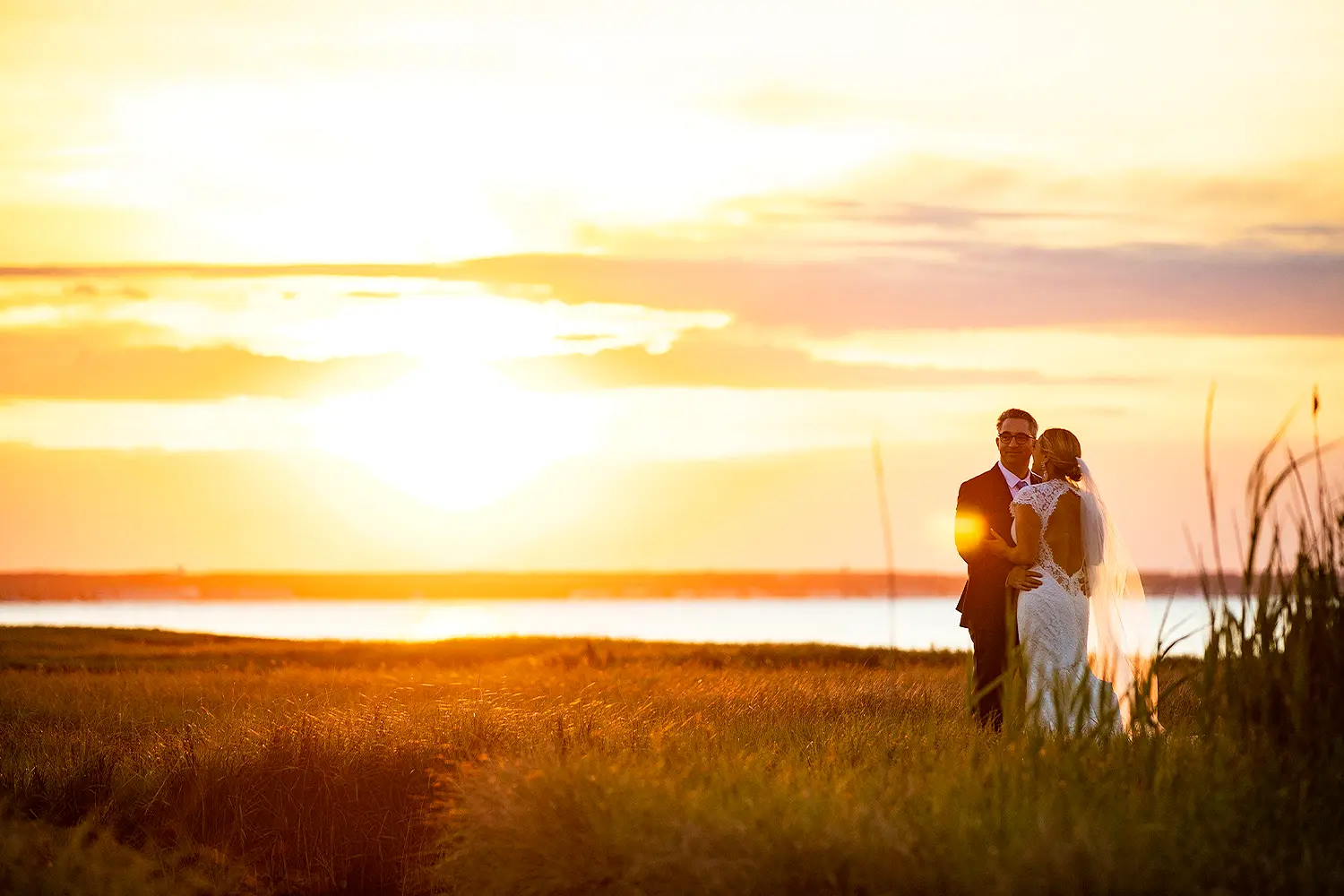 Bride and groom standing together in a grassy marsh at sunset, bathed in golden light with water in the distance.