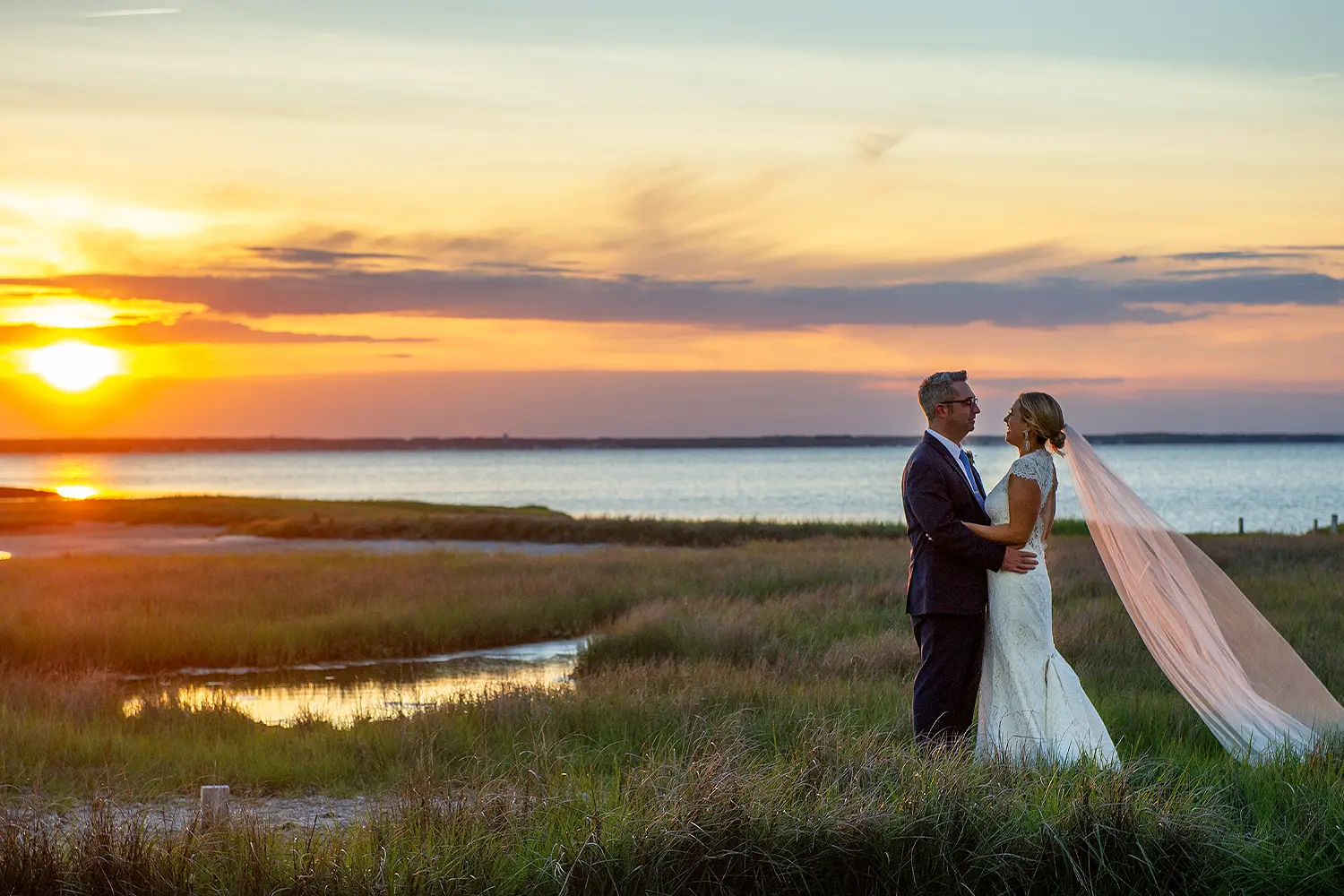 Bride and groom facing each other in a marsh at sunset, with calm water and a colorful sky behind them as the bride’s veil flows in the breeze.