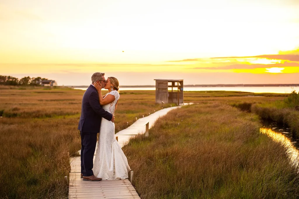 Bride and groom kissing on a wooden boardwalk in a coastal marsh at sunset, with golden light reflecting on the water and grasses.