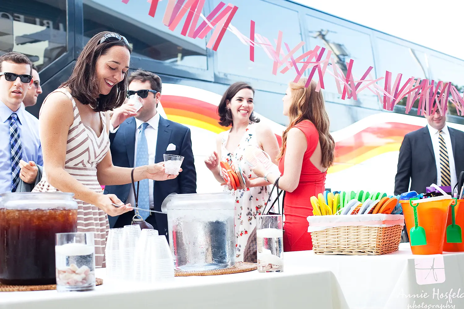 Wedding guests gather around a drink station, pouring beverages and chatting beside a decorated bus with colorful streamers.