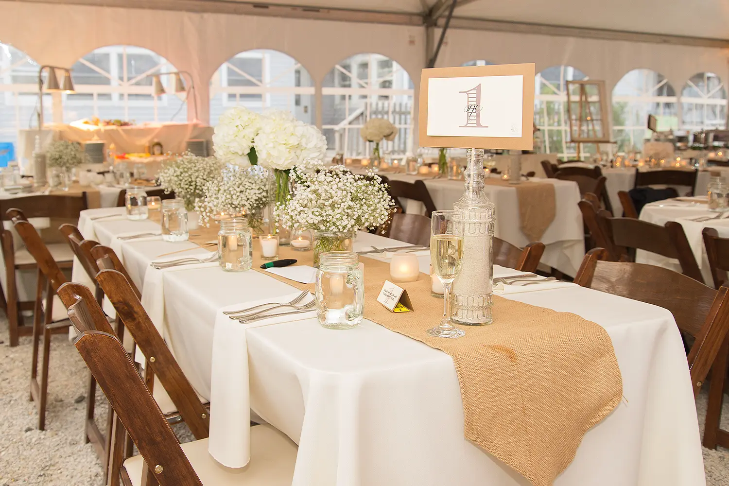Wedding reception table under a tent with white linens, burlap runner, floral centerpieces, candles, and a table number sign.