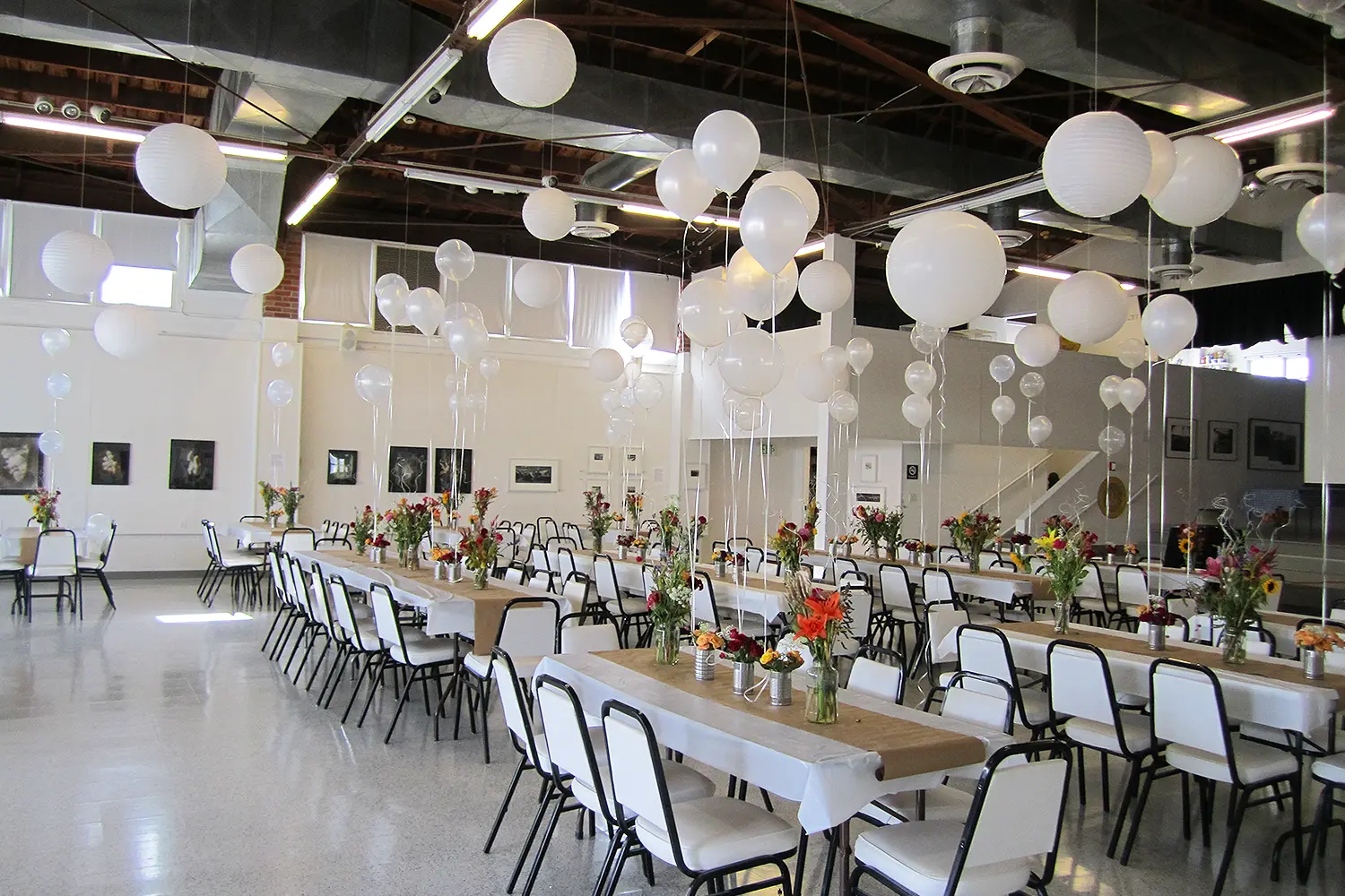 Wedding reception setup inside a bright gallery space with long tables, white balloons, floral centerpieces, and rows of chairs.
