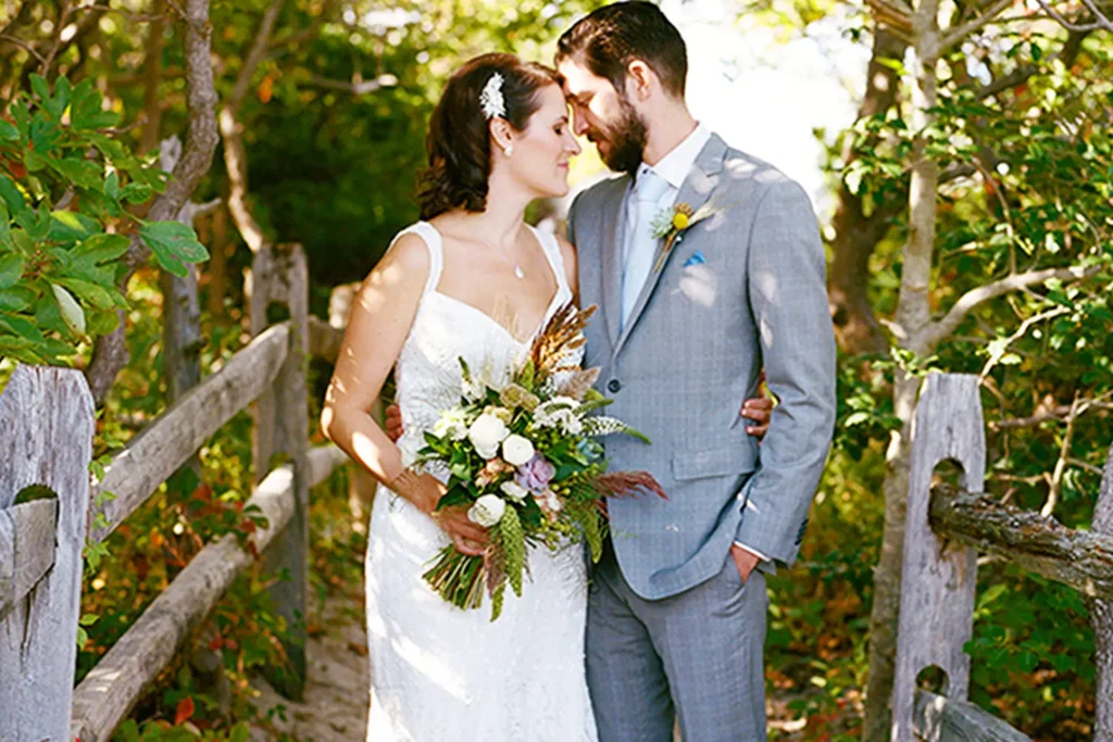 Bride and groom standing closely together on a wooded path, holding a bouquet.