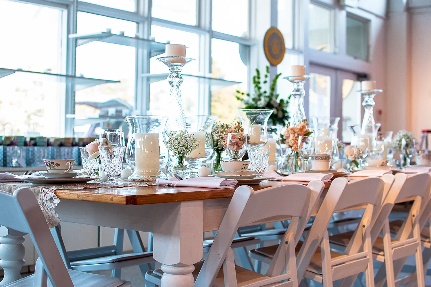 Elegantly set reception table with white chairs, candles, glassware, and soft floral arrangements near large windows.