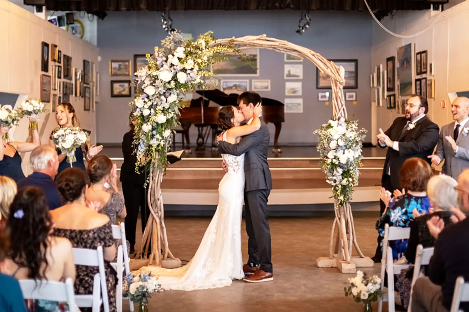 Bride and groom share a kiss beneath a floral ceremony arch as guests applaud indoors.