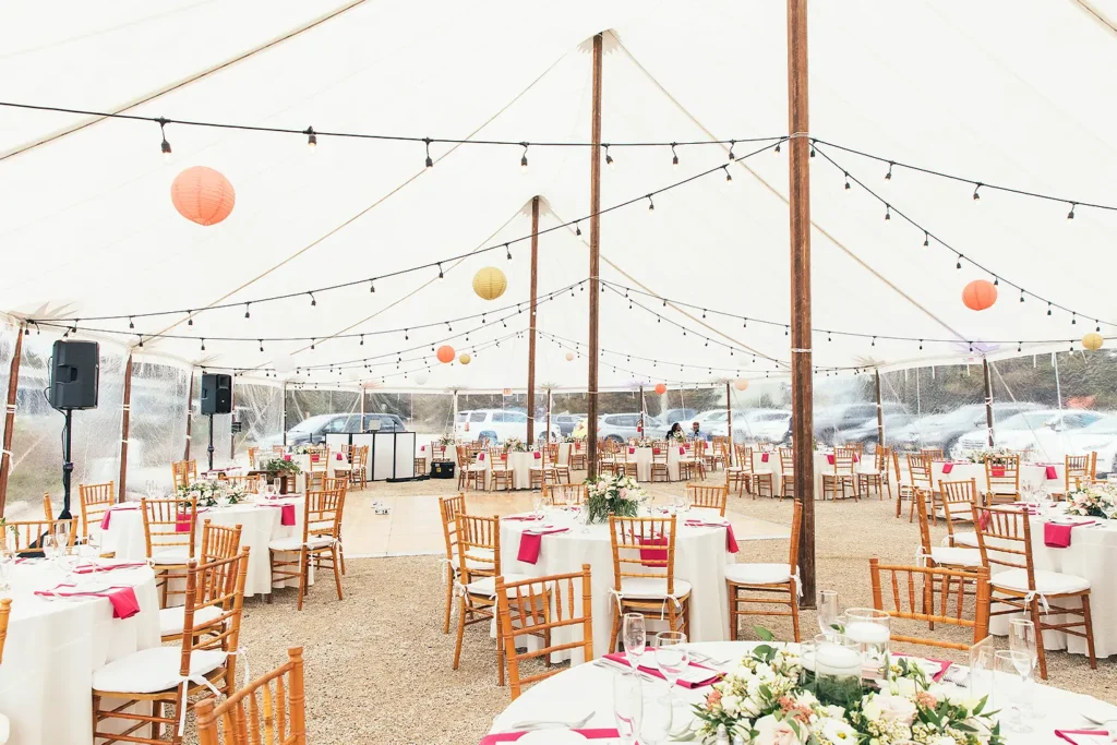 Wedding reception setup inside a white tent with round tables, wooden chairs, and string lights overhead.