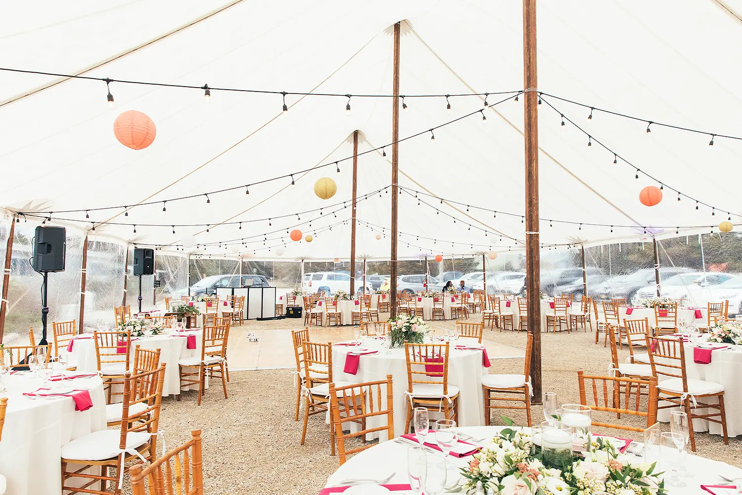Wedding reception setup inside a white tent with round tables, wooden chairs, and string lights overhead.