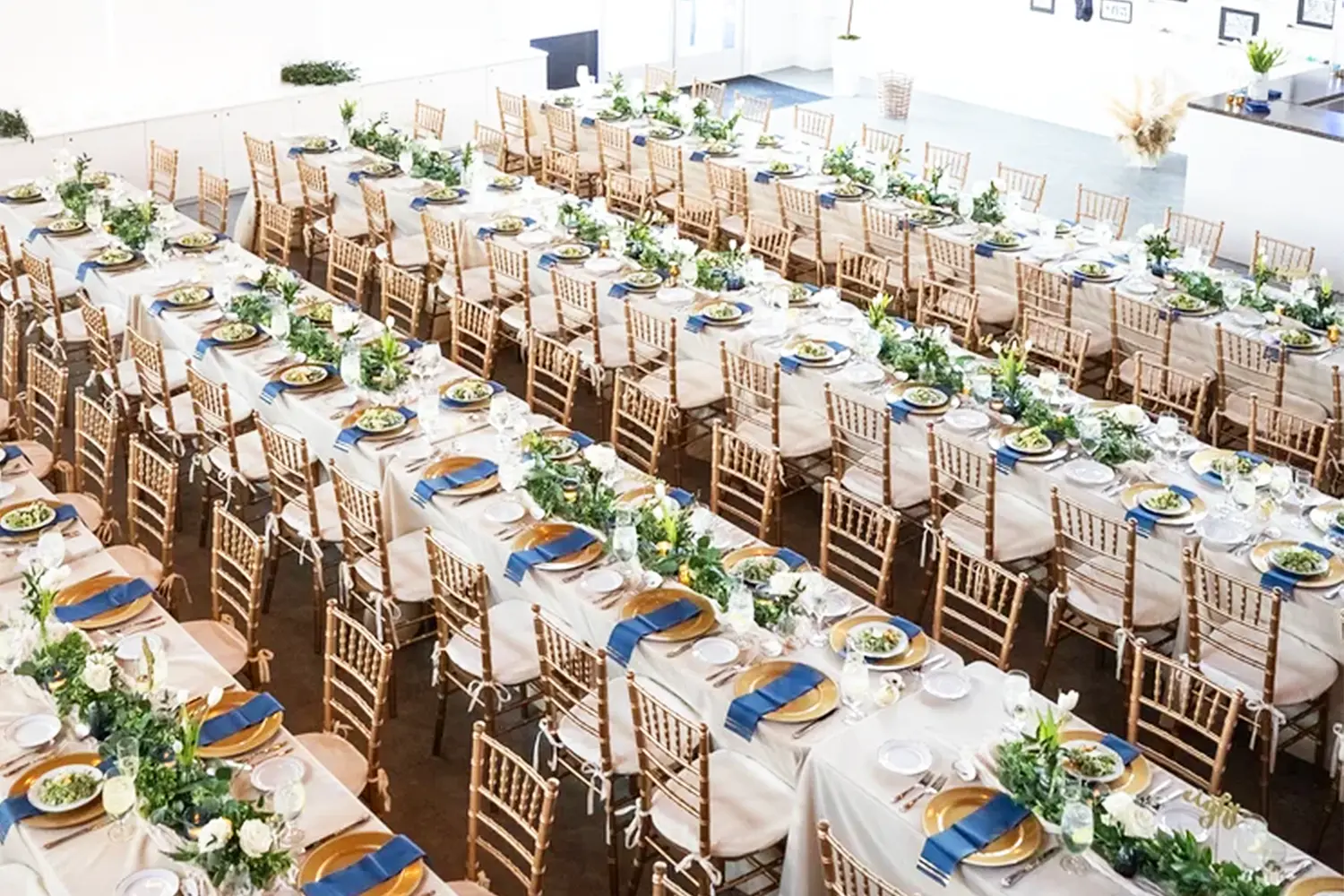 Overhead view of long banquet tables set with white linens, wooden chairs, greenery centerpieces, and gold-rimmed place settings.
