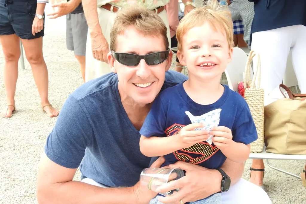 An adult and a young child smile together at an outdoor community event, with the child holding a small handmade paper craft.